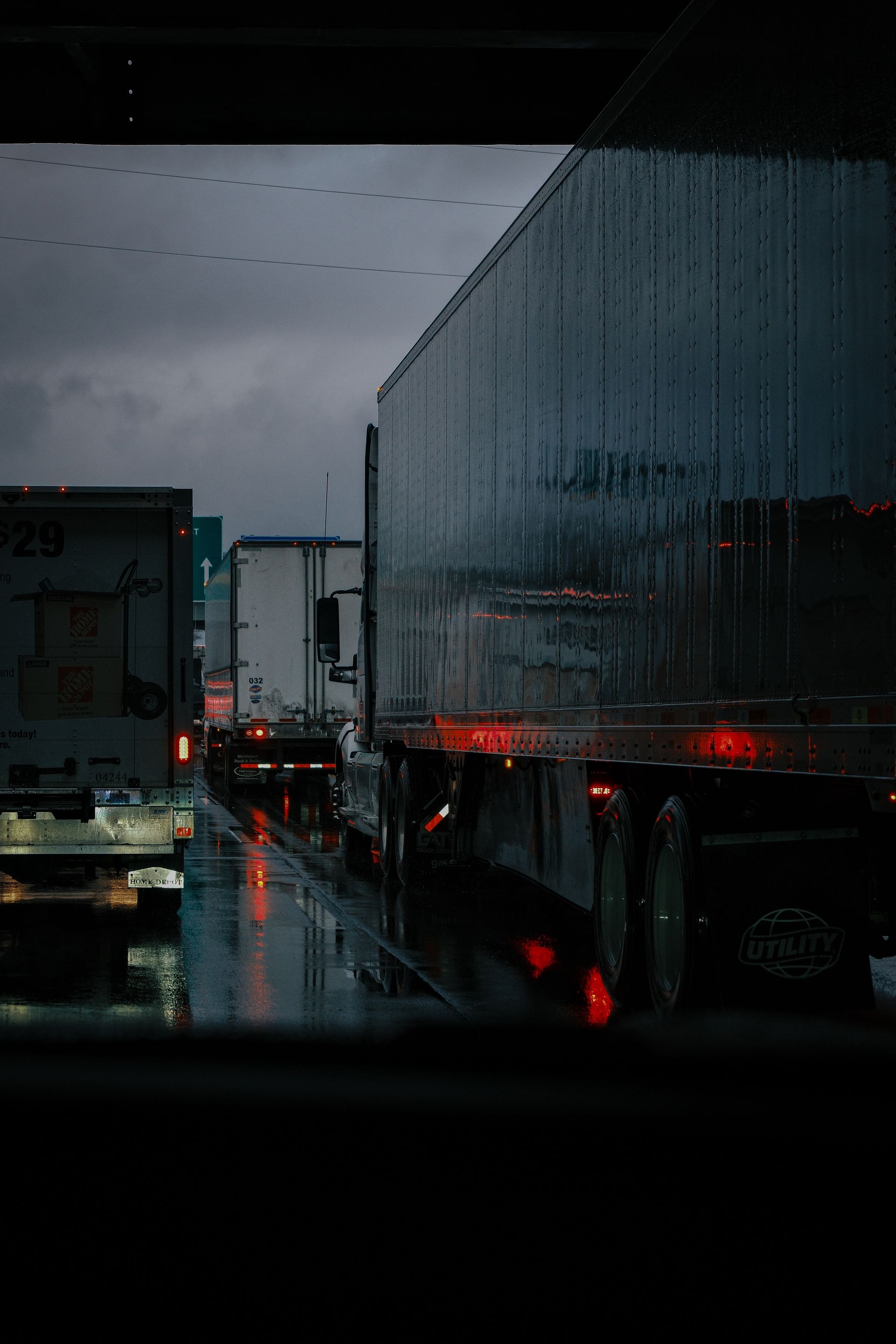 Semi trucks on a wet highway at dusk, red taillights reflecting off the road